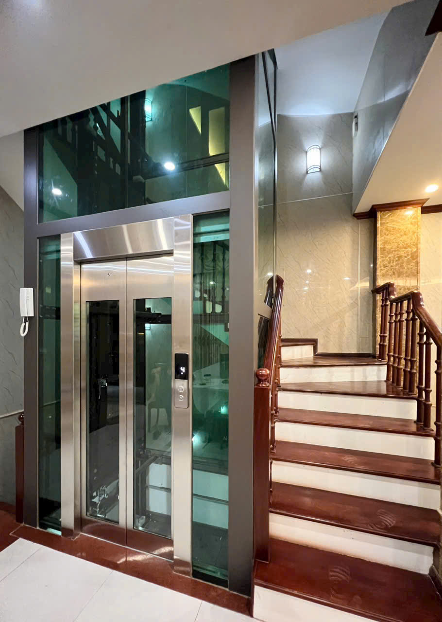 Thang máy gia đình vách kính khung inox lắp cạnh cầu thang gỗ trong nhà sang trọng